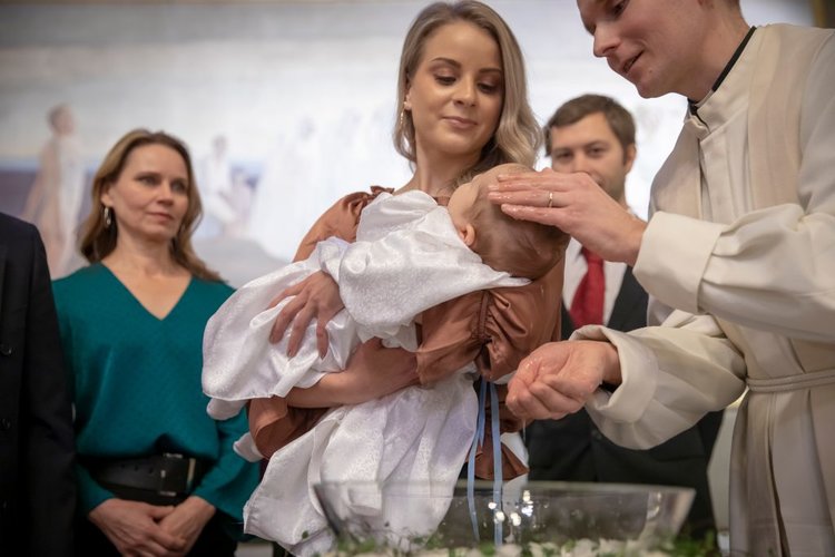 Pappi kastaa lasta, joka on nuoren naisen sylissä. Ympärillä ihmiset seuraa tapahtumia.