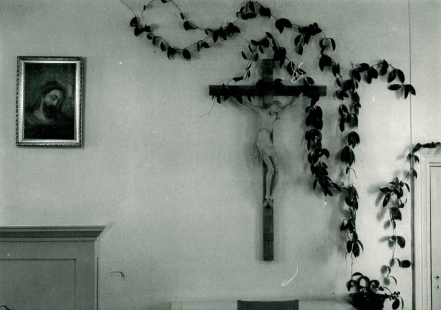 Vnhassa seurakuntasalissa viherkasvi reunustaa krusifiksia ja sen vierellä näkyy Jeesusta esittävä maalaus.