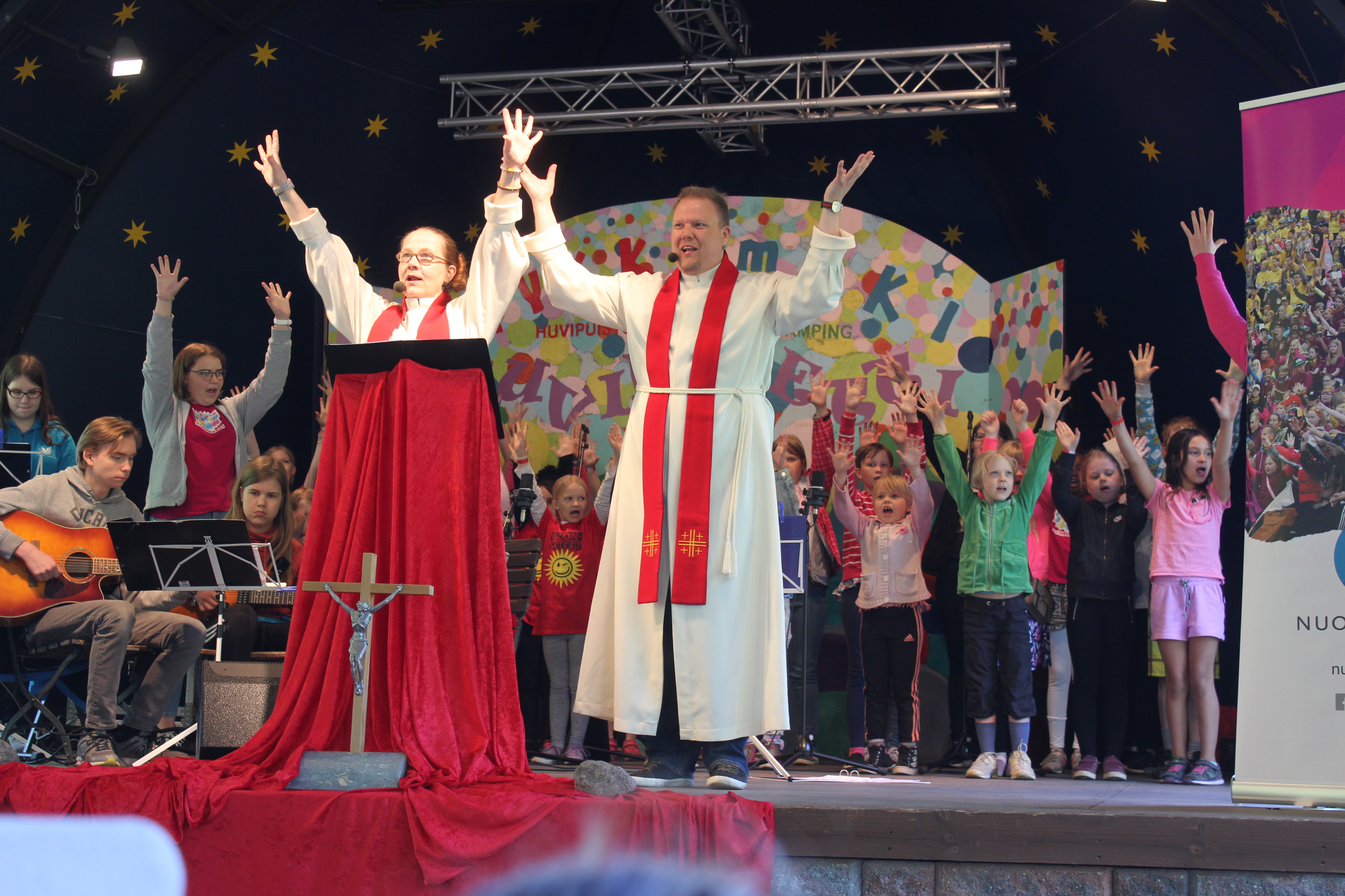Kaksi pappia laulaa kädet kohotettuina, taustalla bändi ja kuorolaisia.