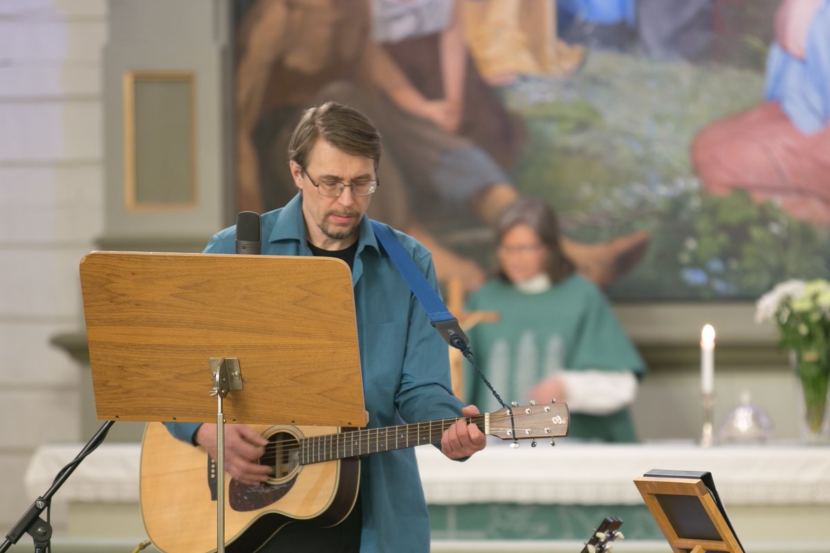 Juha Tanska soittaa kitaraa Jaalan kirkossa. Taustalla Minna Tanska papin asussa kirkon alttarilla. Kuva: K...
