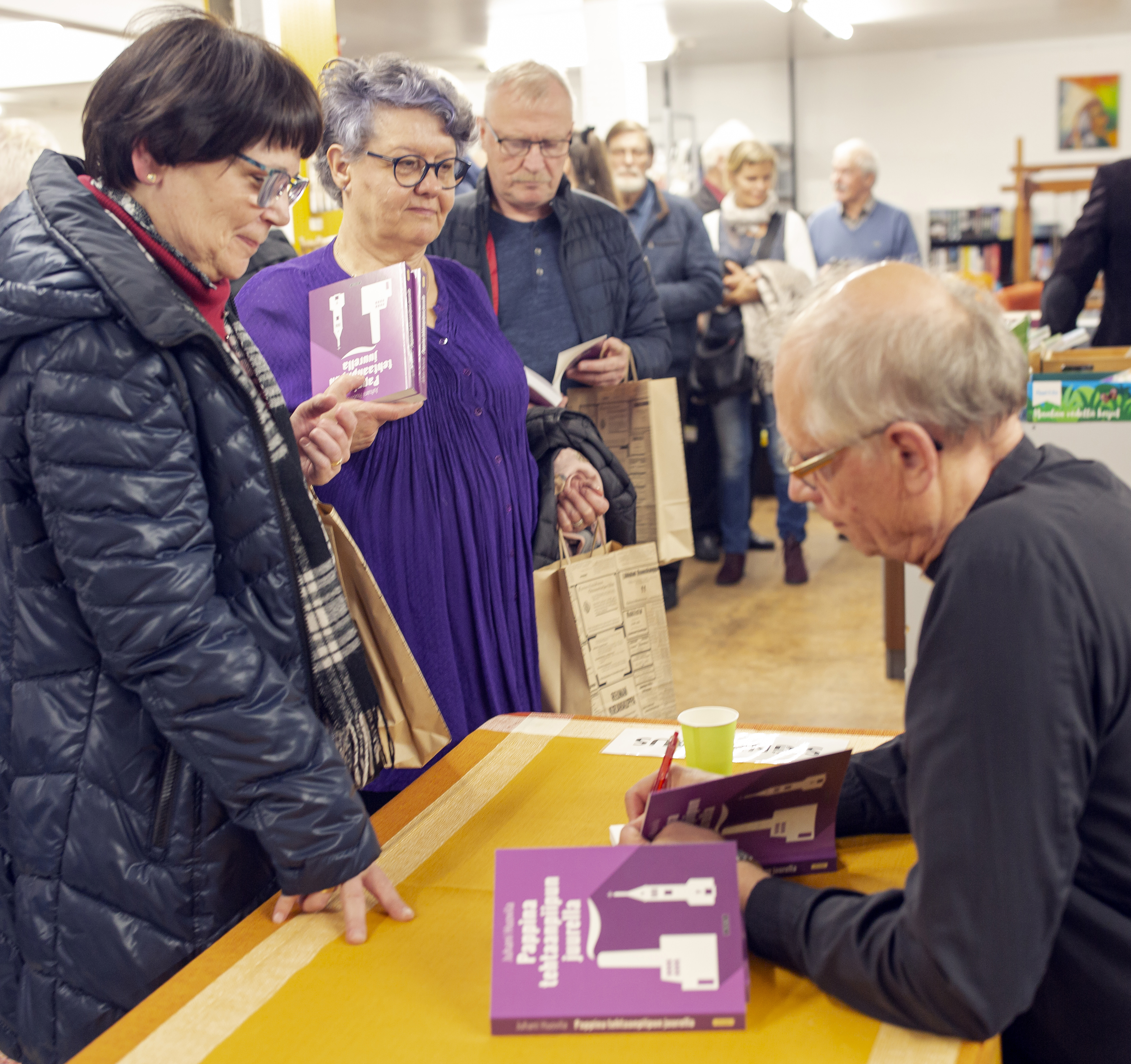 Juhani Huovila signeeraa kirjaansa pöydän ääressä. Signeerausta odottavia henkilöitä seisoo jonossa odottam...