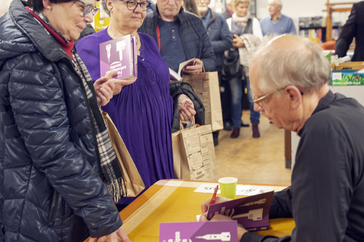 Juhani Huovila signeeraa kirjaansa pöydän ääressä. Signeerausta odottavia henkilöitä seisoo jonossa odottam...