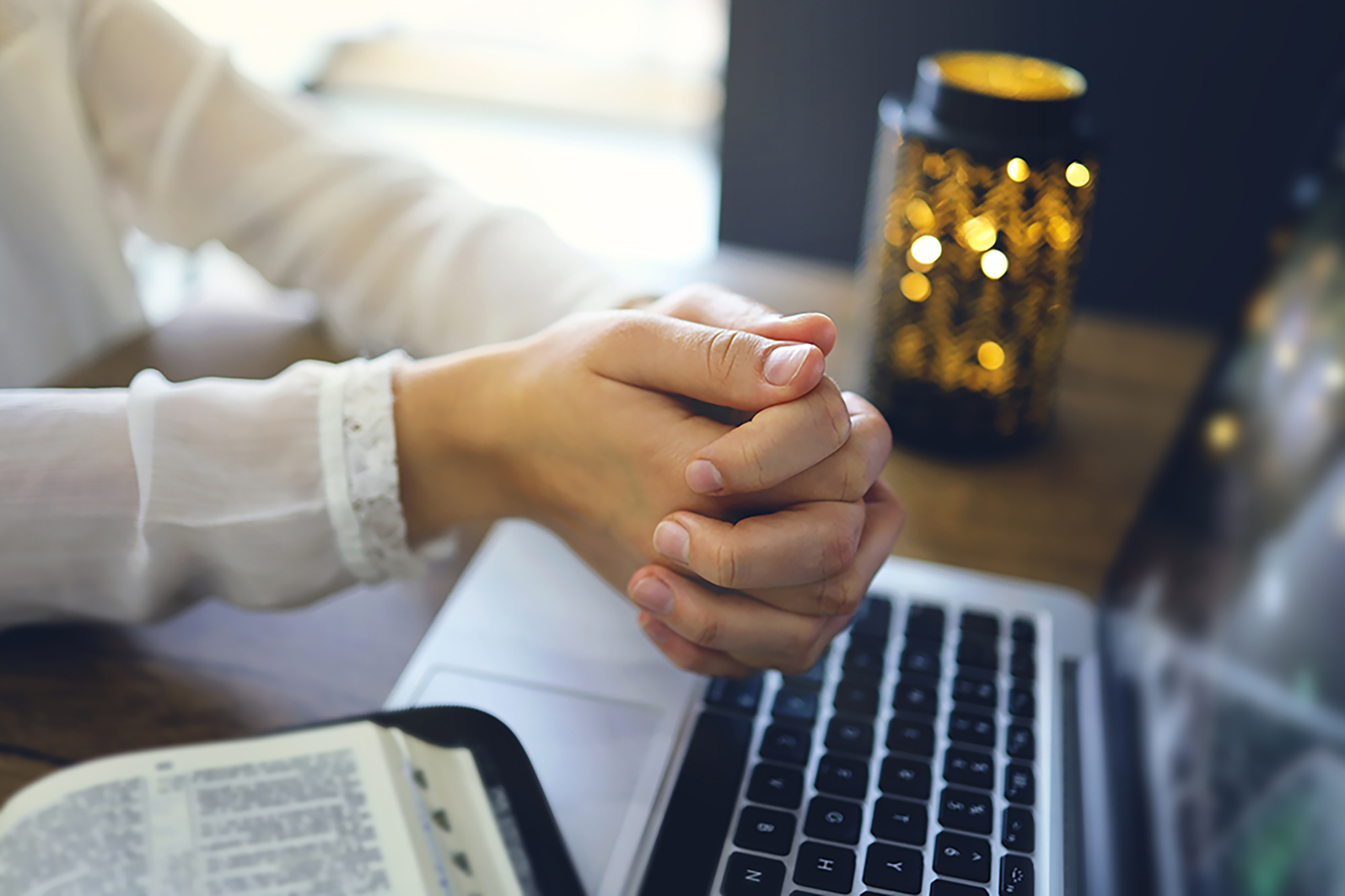 Kuvassa naisen kädet ristissä raamatun päällä ja avoinna olevan kannettavan tietokoneen päällä.