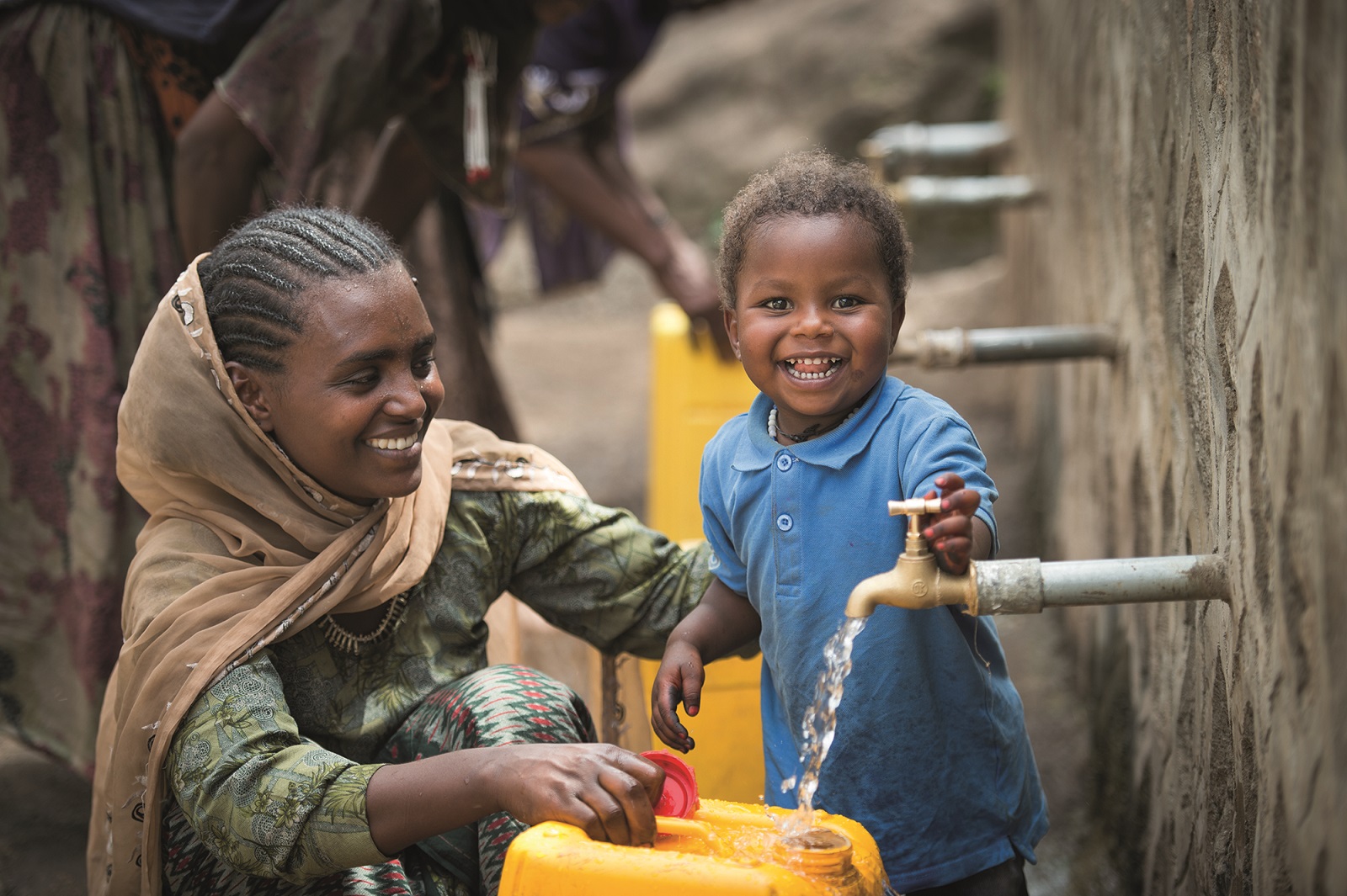 Etiopialainen äiti ja lapsi laskevat hymyillen vesihanasta vettä kanisteriin.
