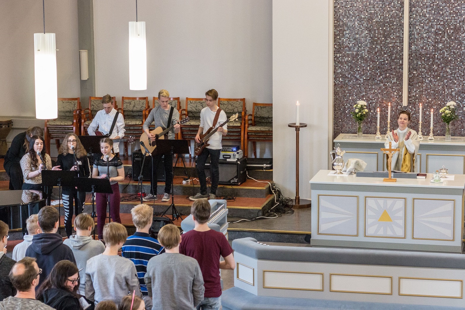 Jumalanpalveluksessa pappi alttarilla, bändi laulaa ja soittaa sekä jumalanpalvelusvieraita.