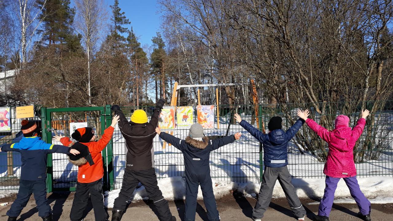 Joukko lapsi seisoo rivissä selkä katsojaa kohti aurinkoisessa kevät säässä Koivistontalon edustalla.