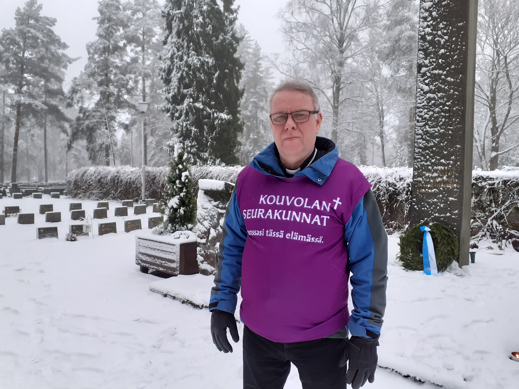 Pappi seisoo lumisella hautausmaalla muistomerkin edessä.