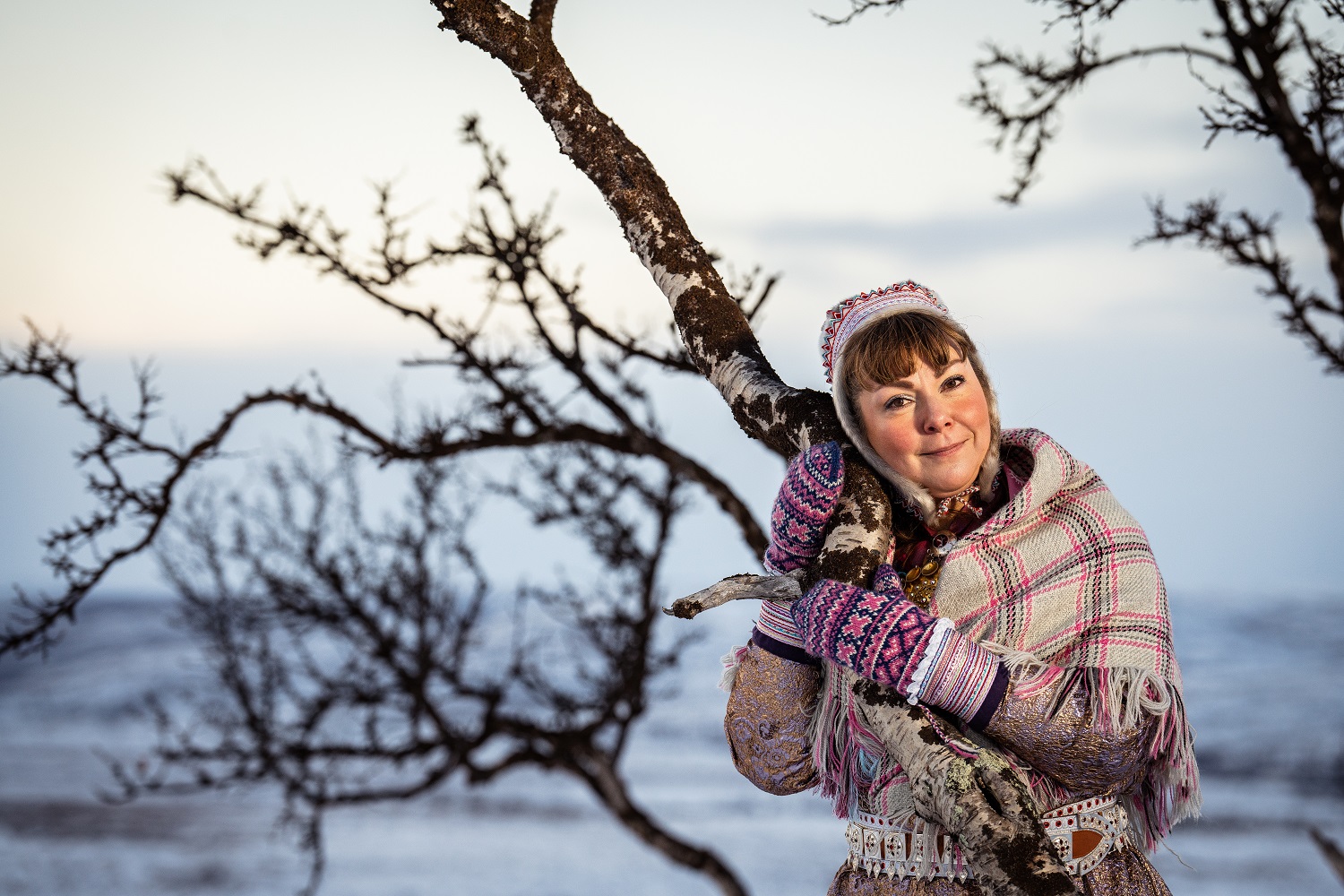 Saamelaispukuun pukeutunut nainen halaa vänkyräistä koivunrunkoa talvikelissä.