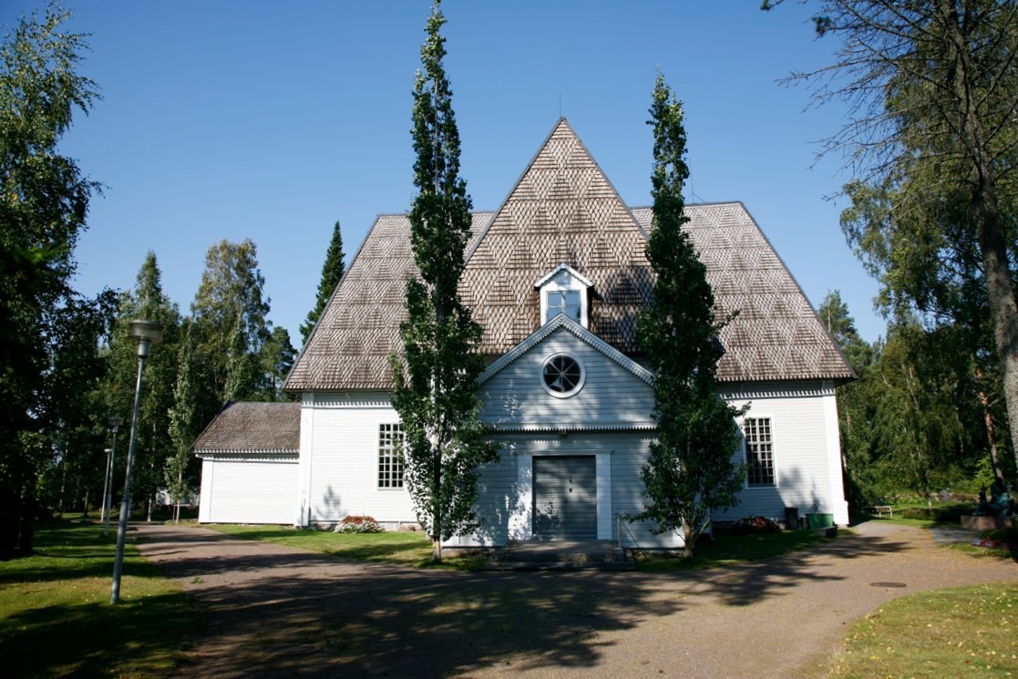 Valkoinen kirkko, jonka sisäänkäynnin molemmilla puolilla kasvaa korkeat kapeat havupuut.