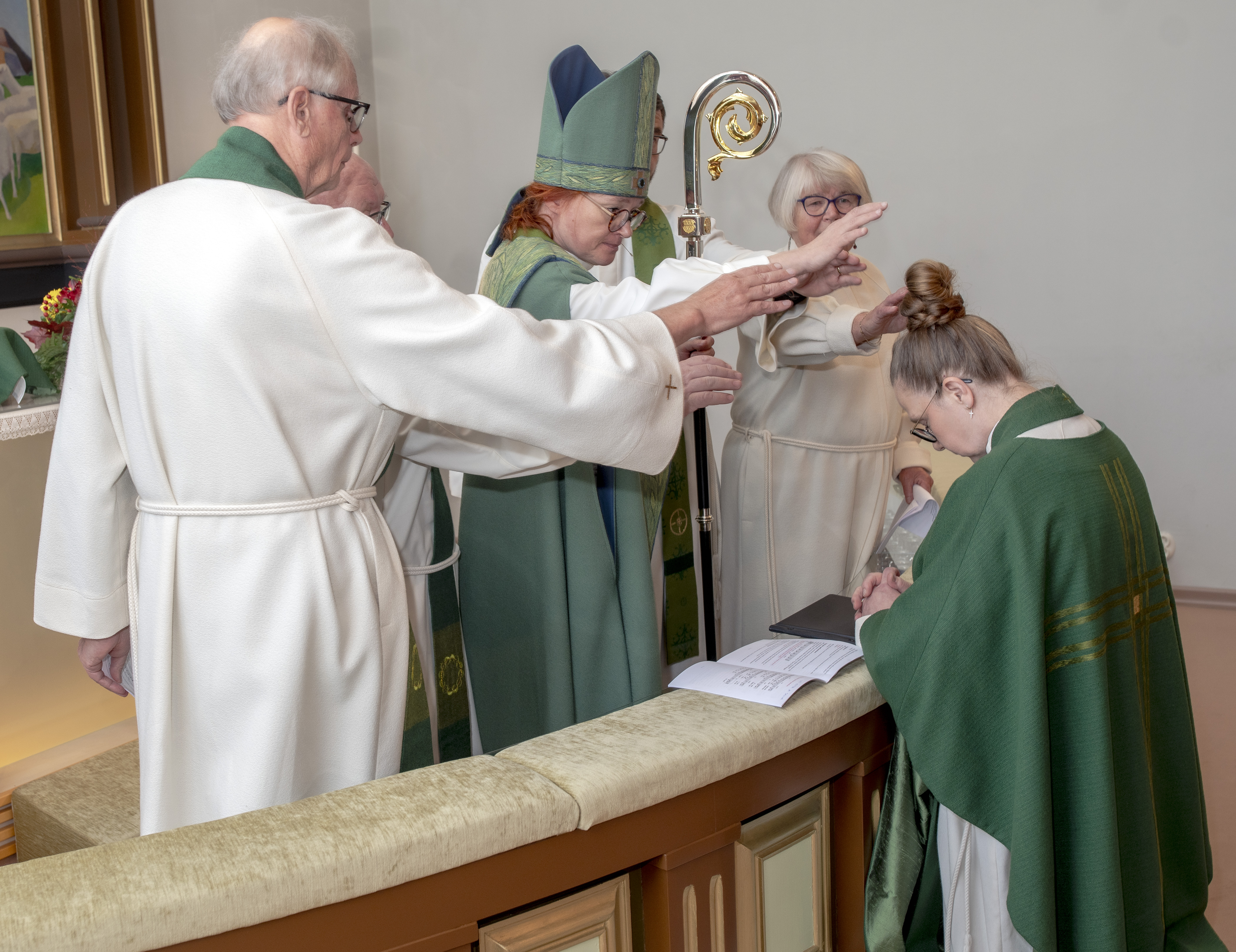 Valkoisiin ja vihreisiin papin alboihin pukeutuneet papit siunaavat kirkon alttarille polvistunutta pappia.