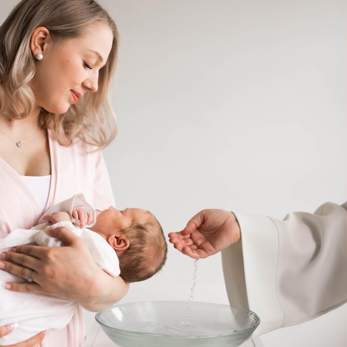 Vaalea-asuinen nainen pitelee sylissään pientä vauvaa, joka on saamassa kastetta.