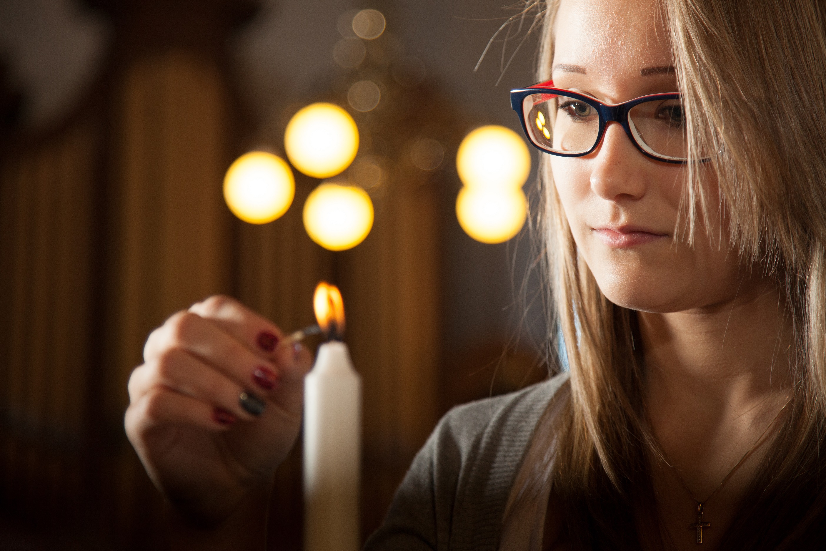 Silmälasipäinen, vaaleahiuksinen nainen sytyttää tulitikulla valkoista kynttilää.