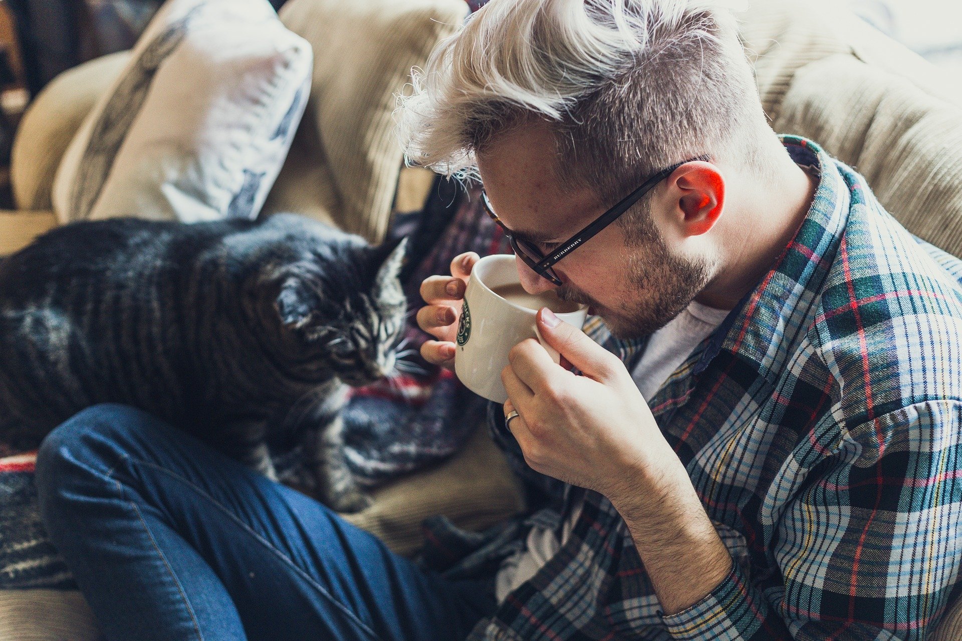Silmälasipäinen mies hörppii kahvia sohvalla kissan vierellä.