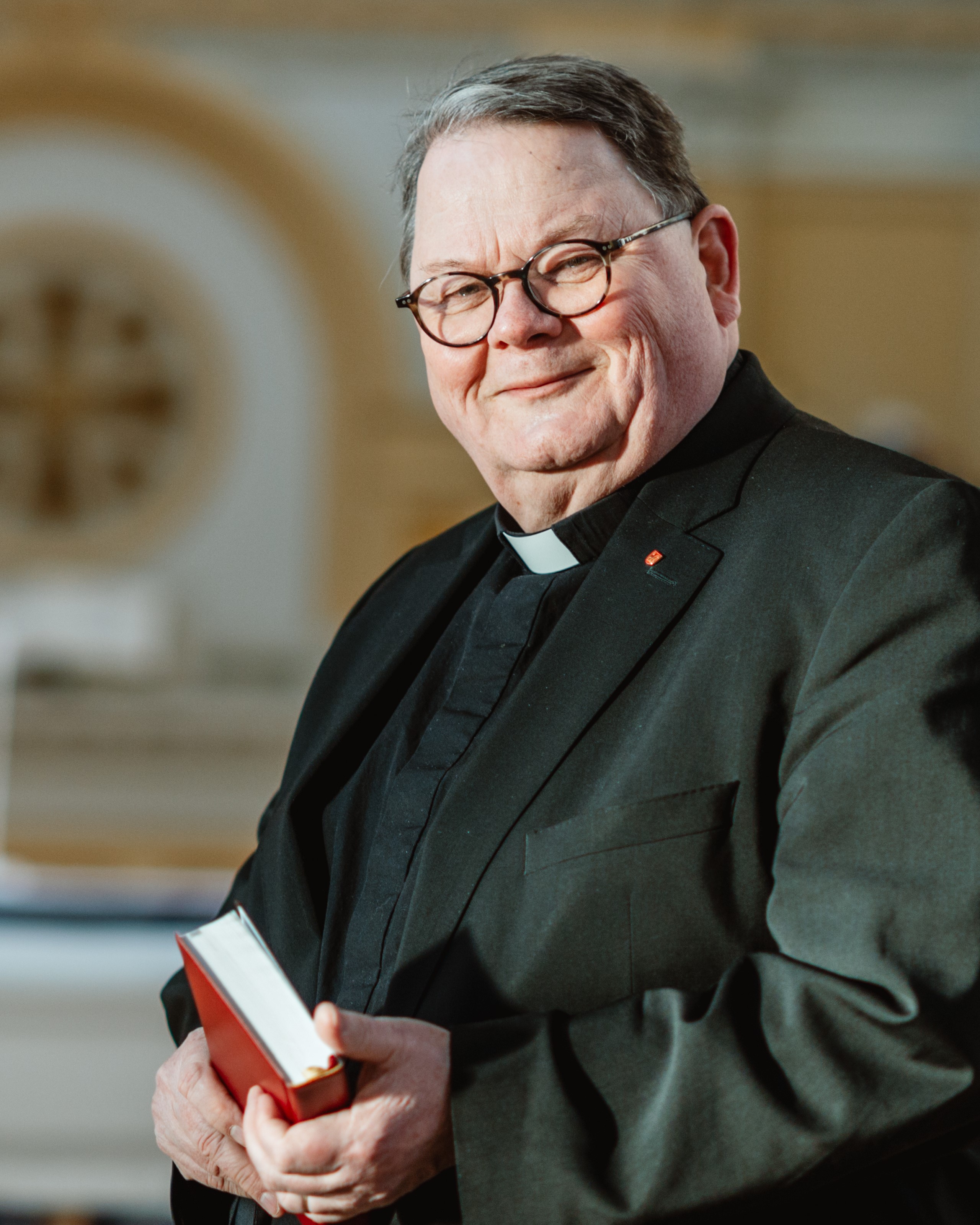 Pekka Huokuna seisoo valoisassa kirkossa papin puvussa kädessään virsikirja.