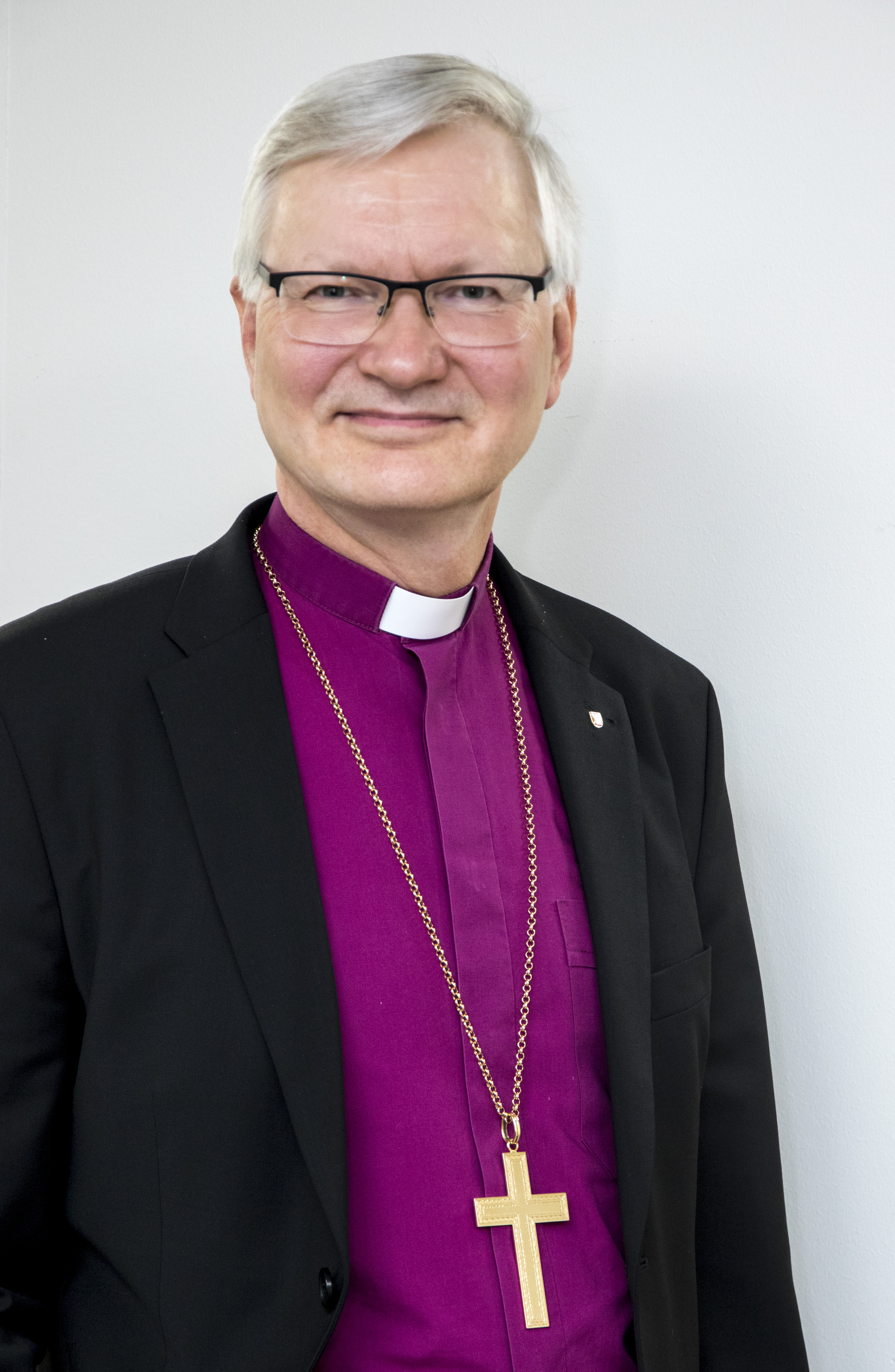 Silmälasipäinen ja harmaahiuksinen piispa viininpunaisessa paidassa ja suuri ristikoru kaulallaan roikkuen.