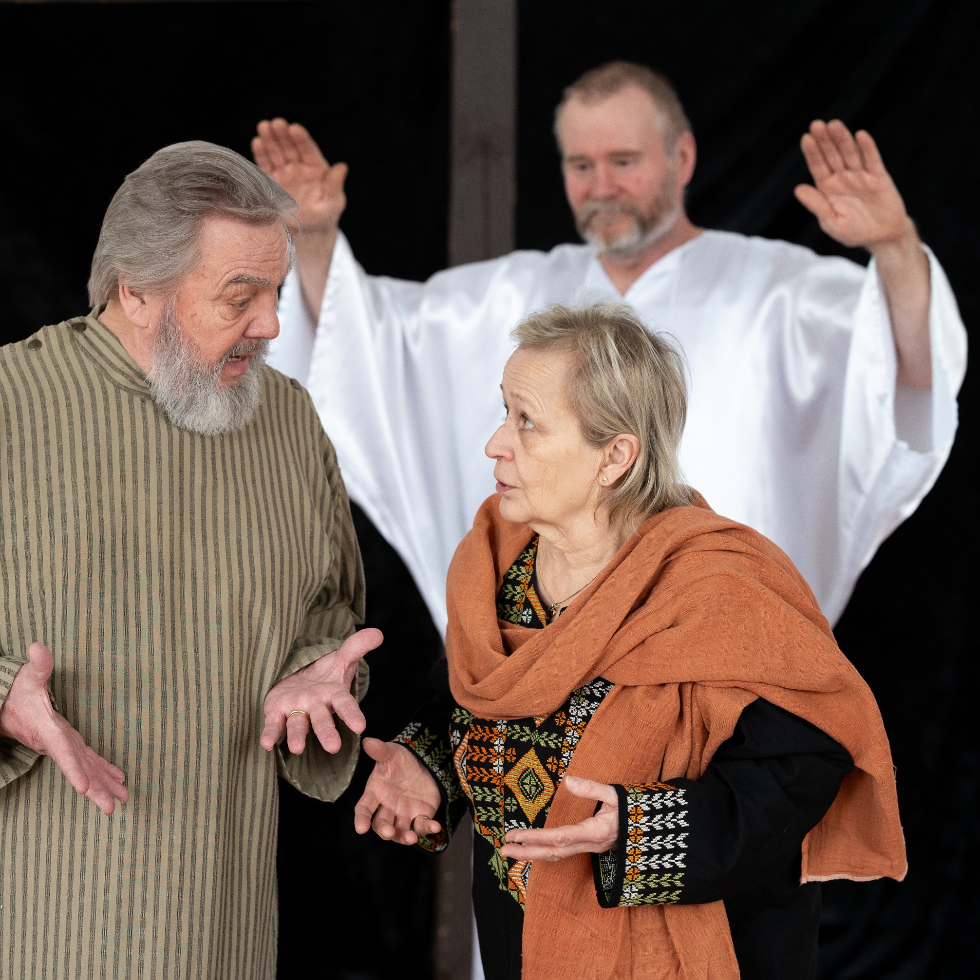 Jeesus on kuvassa taustalla kädet kohotettuina siunaukseen. Kaksi näyttelijää keskustelevat etualalla.