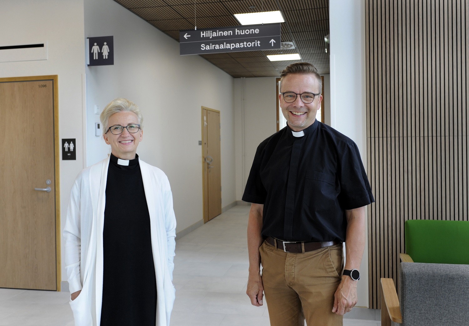 Kaksi hymyilevää pappia sairaalan käytävällä. Opasteessa lukee