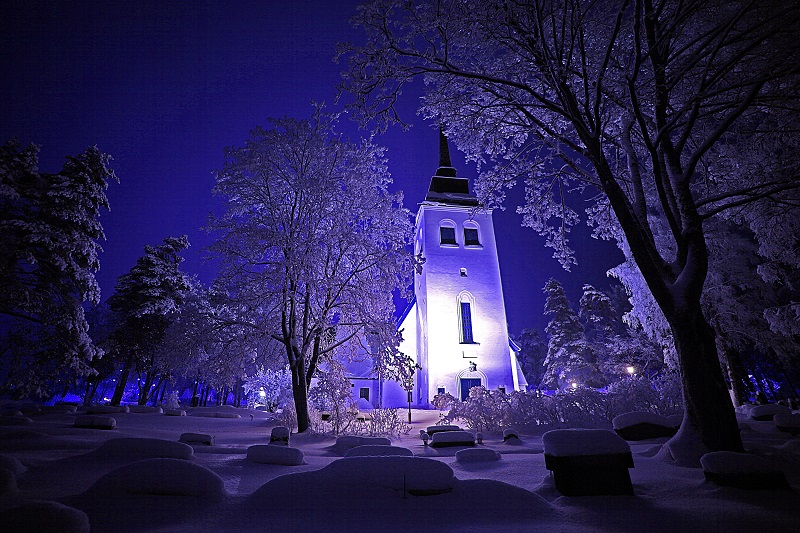 Sinisävyisessä öisessä hämyssä oleva vaalea kirkko talvisen maiseman keskellä.