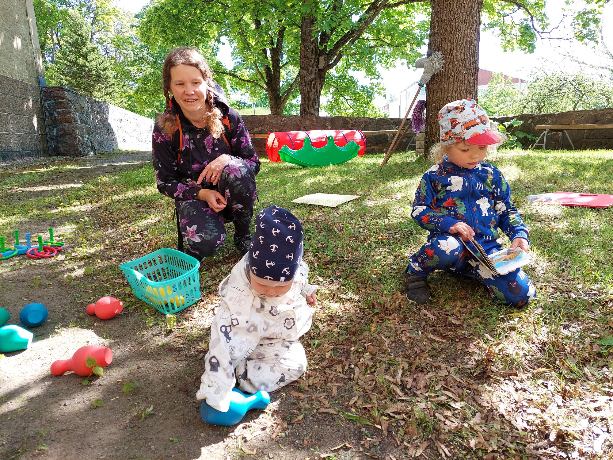 Aikuinen ja kaksi pientä lasta leikkimässä ulkona erilaisilla leluilla.