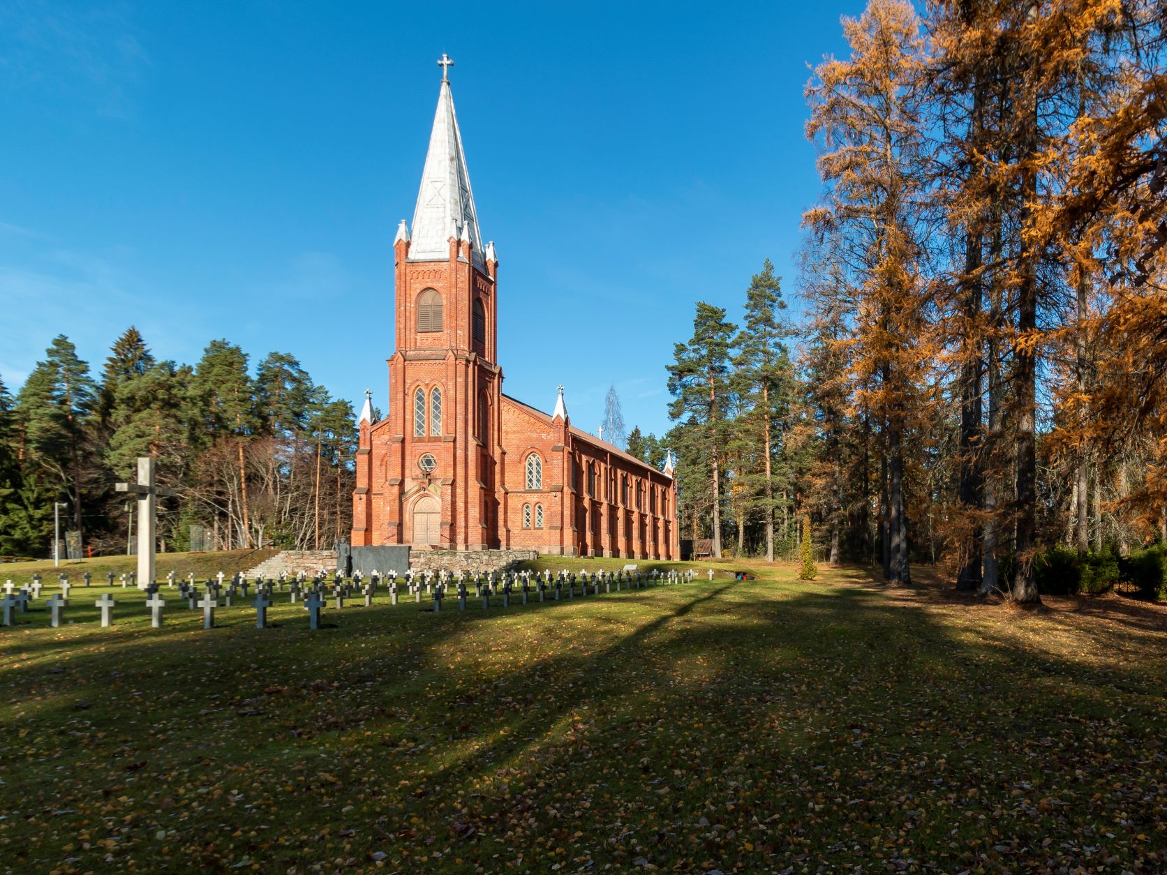 Punatiilinen kirkko aurinkoisessa maisemassa, jossa taivas on lähes pilvetön ja puiden varjot tulevat pitki...