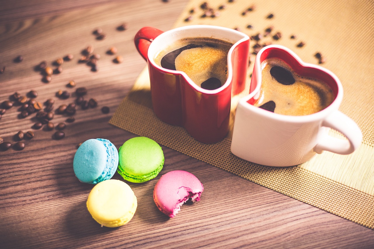 Kaksi sydämenmuotoista kahvikuppia, punainen ja valkoinen. Pöydällä on myös neljä värikästä macaron-leivost...
